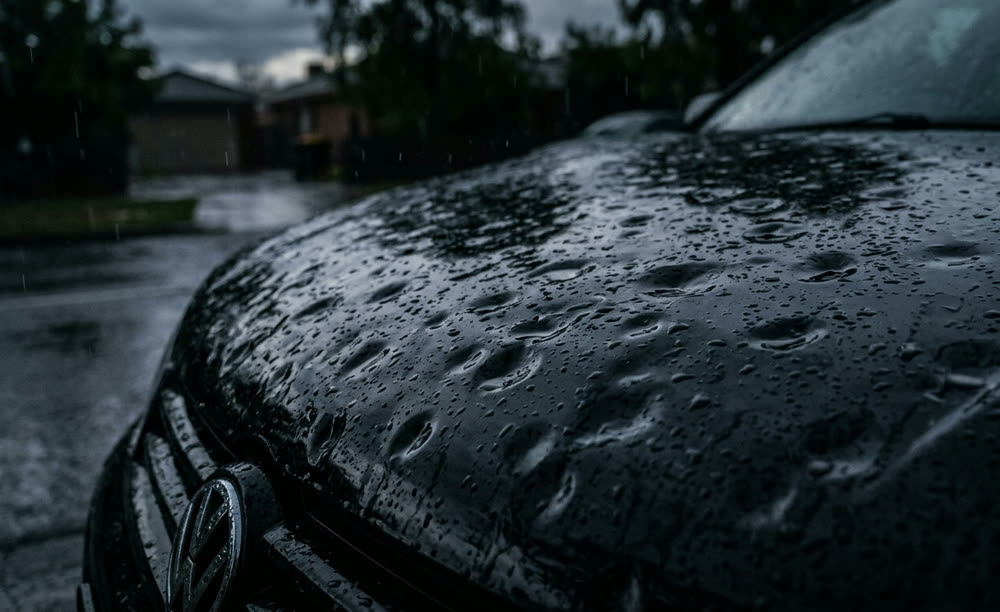 Rain beading across a dark car bonnet dimpled with hail dents